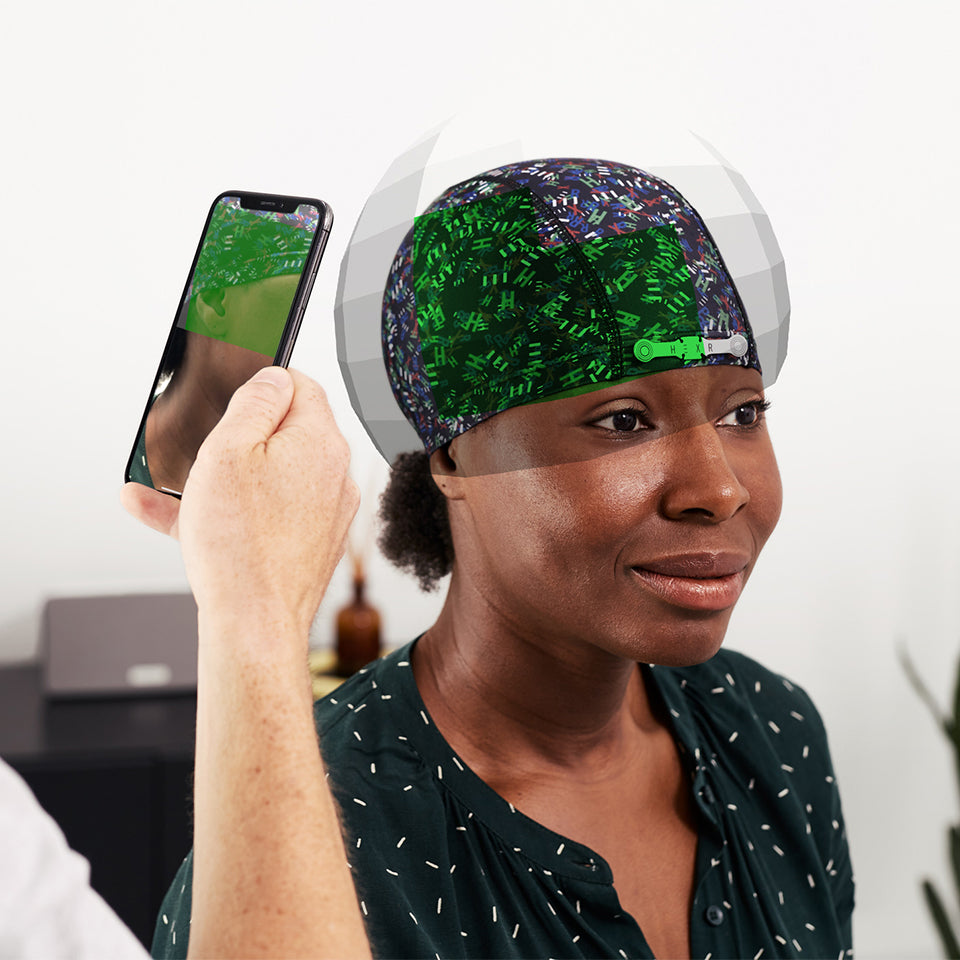 A woman sits wearing the HEXR fitting cap. A hand, holding a smartphone, points toward the lady.